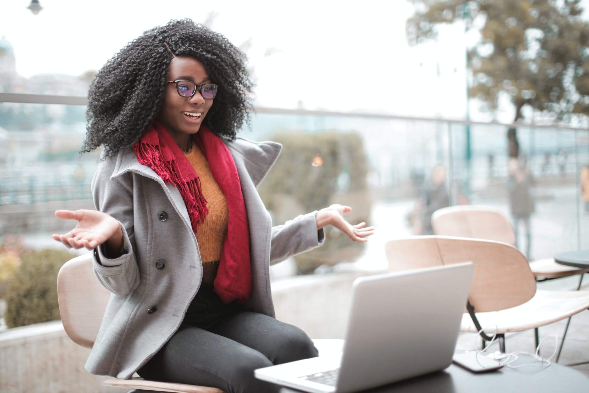 Black woman sitting outside with scarf and coat on looking at MacBook on table