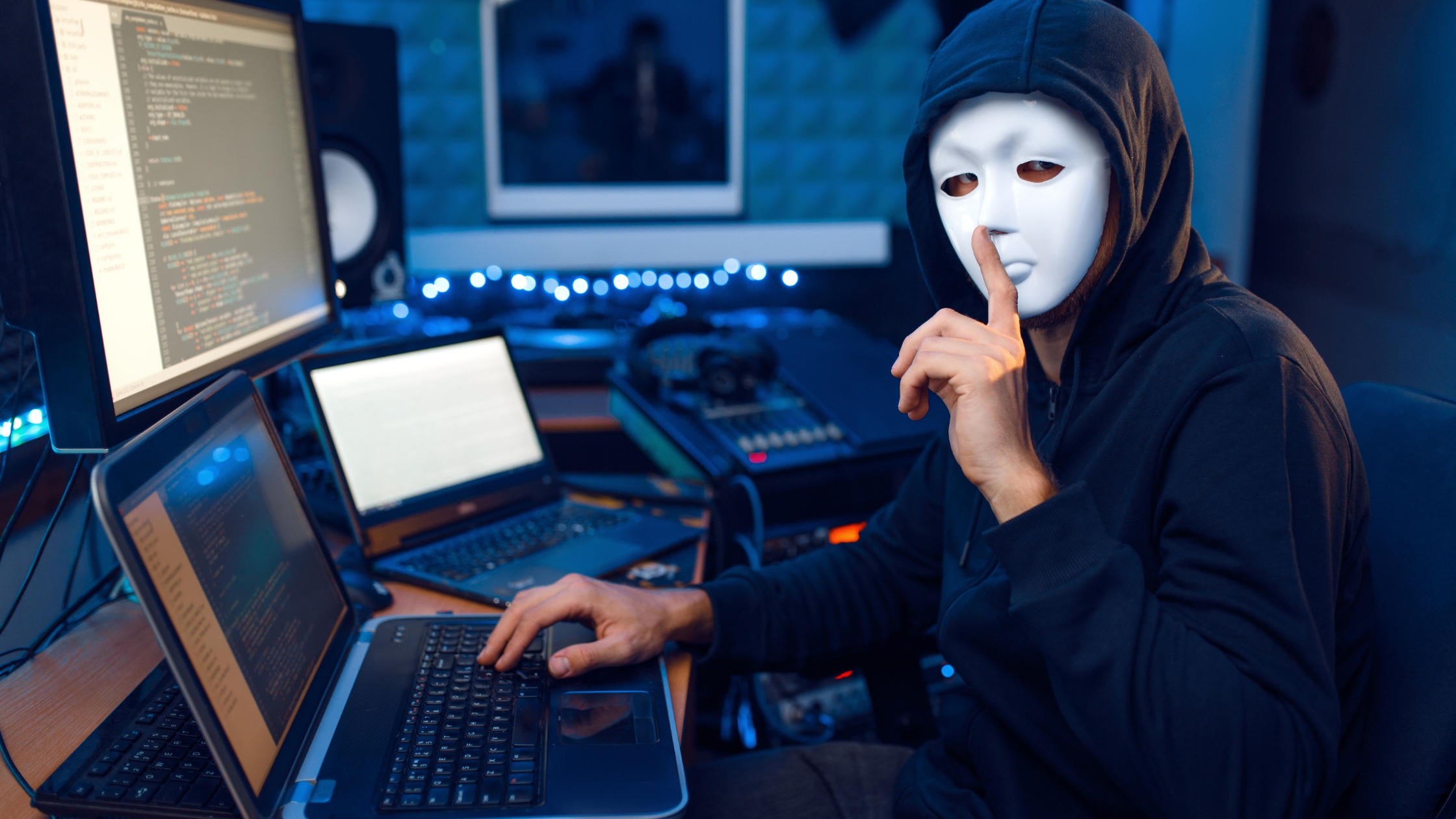 Website hacker wearing white mask and all black at computers on a desk